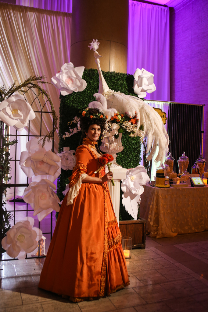 Guest at vanity fair wedding show in orange dress in front of flower and statue installation for venue decor