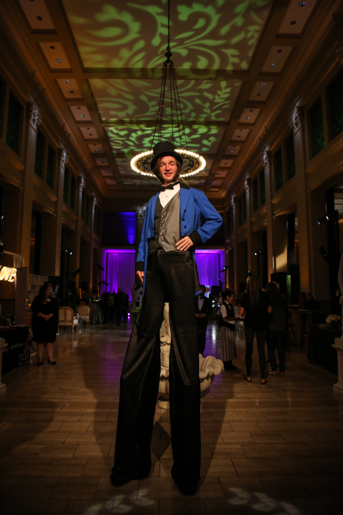 Event performer on stilts underneath green pattern ceiling lighting and chandelier lighting in background