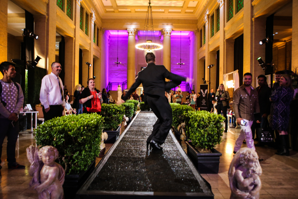 Groom model walking on runway for vanity wedding show with crowd watching, venue decorum and gold and purple wall lighting 