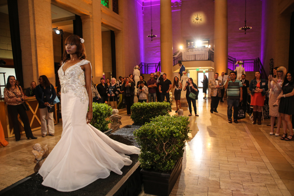 Bride model walking on runway for vanity wedding show with crowd in the background and gold and purple wall lighting around venue