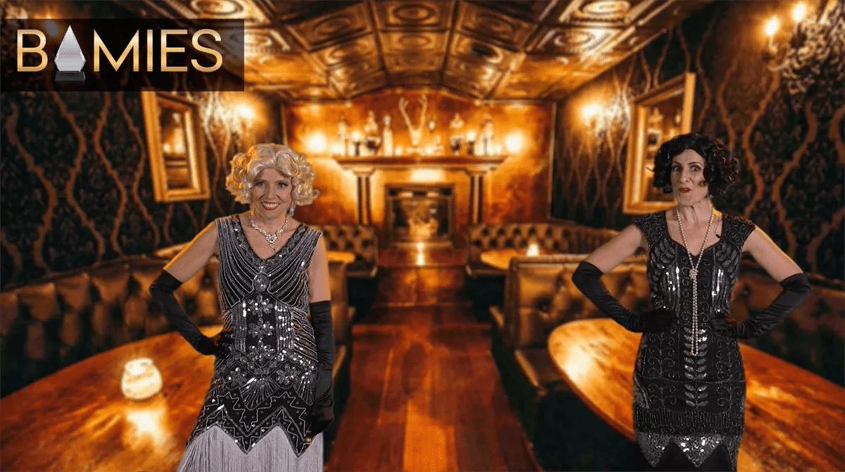 Two characters dressed up in dresses and wigs in front of a lounge background setting