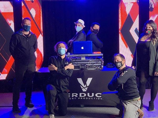 Verducci team posing by the DJ booth with masks on in front of red and black backdrop with blue led lights